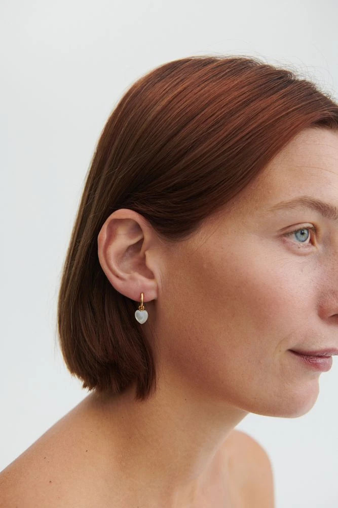 Pernille Corydon Ocean Heart Huggies in Gold being worn by a white woman with dark red hair and blue eyes against a cream background