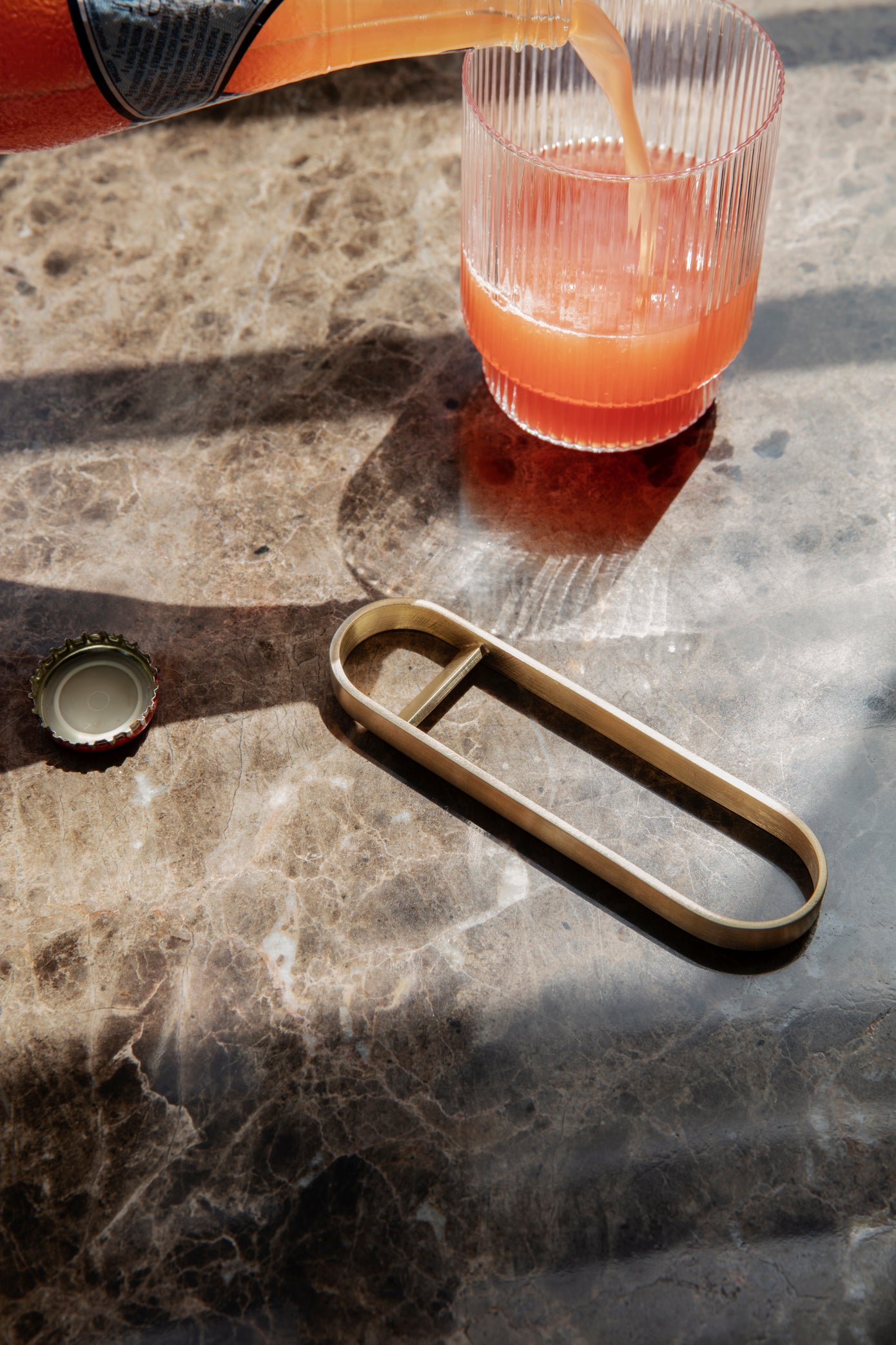 The Fein brass bottle opener lying on a marble tabletop with a ripple glass filled with juice